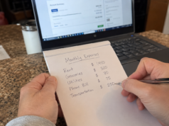 Frugal Living Hacks Ranked by Actual Dollar Impact (Start Here) hands writing a monthly budget breakdown on a notepad next to a laptop showing bank statements
