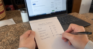 Frugal Living Hacks Ranked by Actual Dollar Impact (Start Here) hands writing a monthly budget breakdown on a notepad next to a laptop showing bank statements