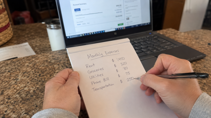 Frugal Living Hacks Ranked by Actual Dollar Impact (Start Here) hands writing a monthly budget breakdown on a notepad next to a laptop showing bank statements
